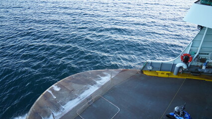 A ferry departs from the port, navigating the serene waters of the ocean. The surface of the water is calm, while the ship's deck is visible, featuring safety equipment and ropes neatly arranged.