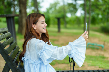 Naklejka premium Asian woman in blue blouse and white pants sitting on black bench, taking a selfie with phone, smiling. Background includes trees and grass in park, capturing cheerful, fun moment outdoors