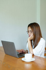 Asian woman in glasses and white shirt working on a laptop at a wooden table, hand on chin. Background includes a coffee cup. Captures focused, professional moment of remote work.