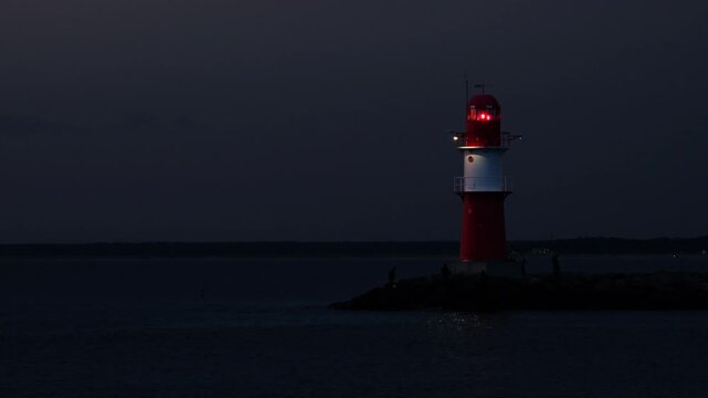 Red light beams of the lighthouse in Warnem&uuml;nde, Germany. Objects of water transportation, tourism, navigation on the Baltic Sea at night concept. People fishing, walking and biking around the tower.