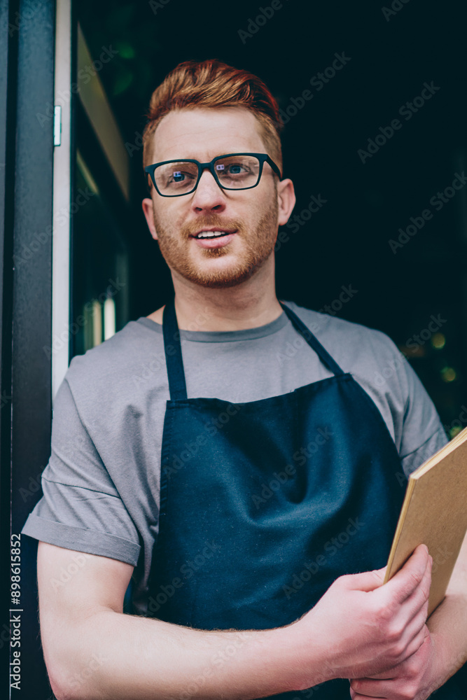 Poster professional caucasian waiter in black apron holding menu and smiling while inviting guests to own b