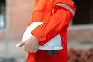 An engineer or foreman in orange coverall uniform is holding a white safety helmet or hardhat,...