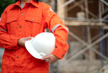 An engineer or foreman in orange coverall uniform is holding a white safety helmet or hardhat,...