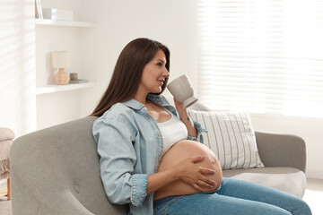 Beautiful pregnant woman with cup on sofa at home