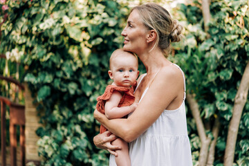 Mom holds her baby for a walk along a green street on a hot summer day.