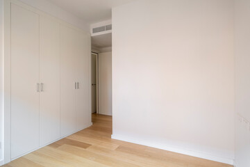 A minimalist bedroom featuring a built-in wardrobe with white doors, natural wood floor, and a wall ventilation system creating a modern and clean look.