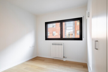 This bright empty room features large windows allowing ample natural light, wooden floors, white walls, and a radiator, providing a fresh and inviting space.