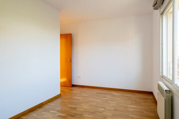 Spacious empty room with a beautiful wooden floor and ample natural light from a large window, featuring a simple white wall and a warm wooden door slightly ajar.