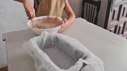 Person whisking brownie batter in a white bowl near a prepared baking pan