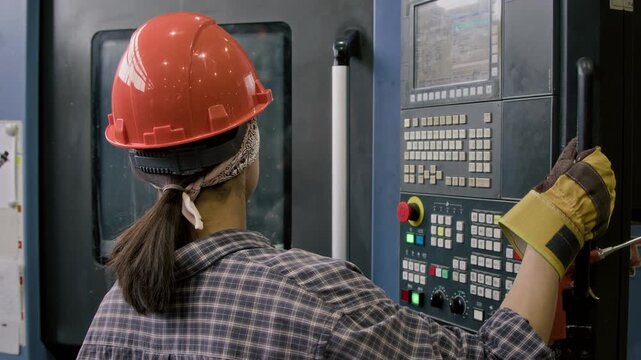 Back view of workwoman in orange safety hardhat controlling production processes in factory, coming up to industrial control panel and moving it by handle