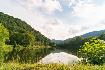 Mountain lake with beautiful reflection. Panoramic landscape with cloudy sky, hills and forest.