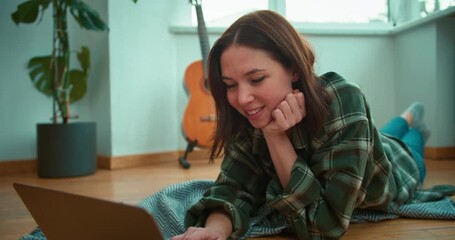 Happy brunette girl lies on the floor and writes down her ideas on a gray laptop near a guitar in a modern apartment