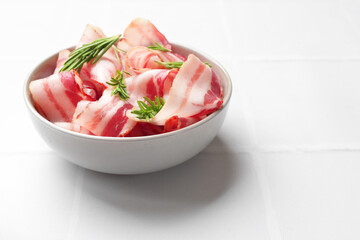Slices of raw bacon and rosemary in bowl on white tiled table, space for text