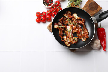 Delicious bacon slices in frying pan and products on white tiled table, flat lay. Space for text