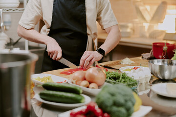 Unrecognizable male hand holding sharp knife and slicing fresh bell pepper on wooden cutting board, he surrounded by kitchenware and vegetables