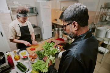 High angle shot of chef sorting fresh lettuce while his sous chef cutting tomatoes in blurred background