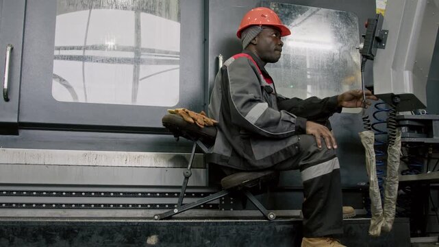 Side full shot of Black male technician in protective coveralls and hardhat sitting by industrial controller and operating automated machinery production process in manufacturing plant