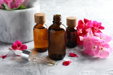 Geranium essential oil in bottles, pipette and beautiful flowers on light grey textured table