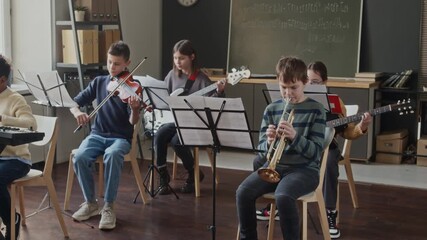 Five multi-ethnic children sitting in front of music stands while playing musical instruments in classroom during lesson - Powered by Adobe