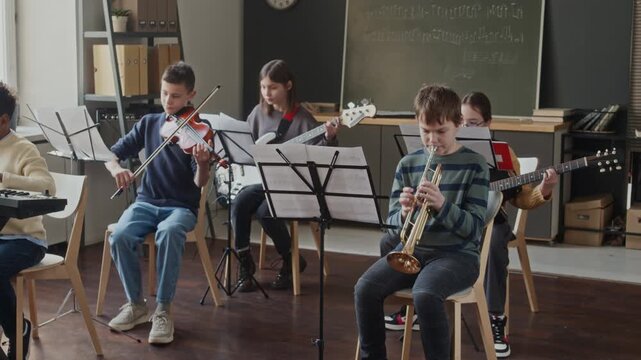 Five multi-ethnic children sitting in front of music stands while playing musical instruments in classroom during lesson