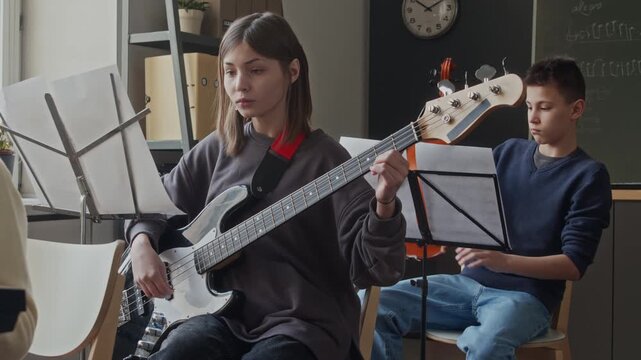 Teenager girl playing electric guitar looking at sheet music in classroom, boy with violin in background
