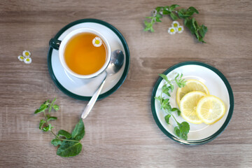 Chamomile Tea with Lemon Slices. Overhead view of a cup of chamomile tea with a small flower floating in it, next to a plate of lemon slices and fresh mint leaves on a wooden table.