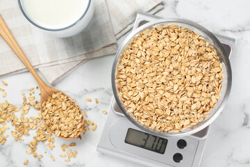 Kitchen scale with bowl of dry oats, milk and spoon on white marble table, flat lay