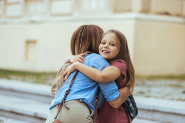 A smiling young girl with a backpack receives a warm hug from her mother, evoking a sense of care...