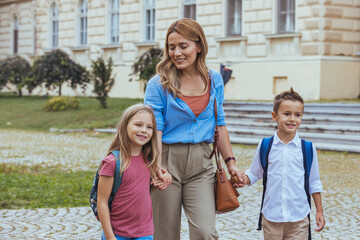 A cheerful Caucasian woman walks her young daughter and son to school, the children carry backpacks and wear casual clothing, with a school building in the background.