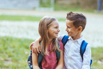 Smiling Caucasian boy and girl carry backpacks standing before a school, exuding eager anticipation for a day of learning and play.