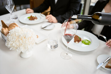 Couple having lunch in restaurant while waiter pouring red wine into glass