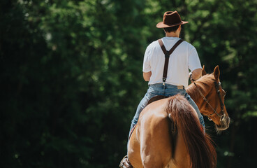 Man wearing suspenders and cowboy hat riding a horse outdoors in a rural setting with green trees...