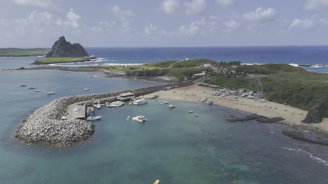 Drone flies to the right over bay with port and Praia do Porto Santo Antonio and Ilha Sela Gineta in background in Fernando de Noronha, Pernambuco, Brazil