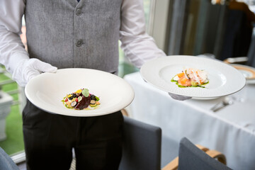 Cropped photo of male waiter in uniform carrying two plates of salads in a restaurant