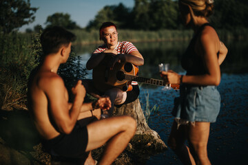 Group of friends spending a summer day outdoors by the lake, playing guitar and relaxing in nature.