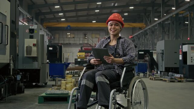 Full portrait of happy Latin female manager with disability sitting in wheelchair, wearing protective uniform and hardhat, posing in plant workshop with tablet and looking at camera