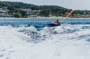 Naklejka premium Man enjoys an adventurous summer activity of tubing behind a speedboat on the sea, with a scenic coastal background.