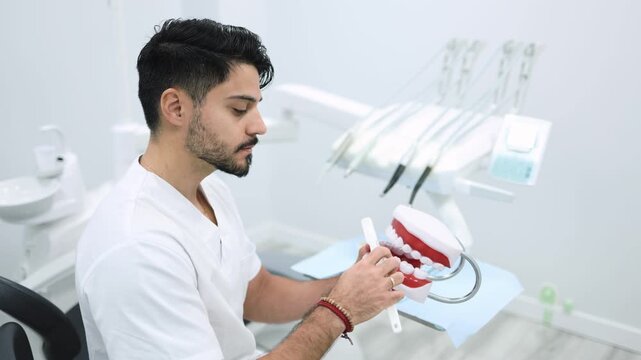 Dentist demonstrating tooth brushing with jaw model in modern dental clinic. Zoom in of Hispanic dentist scrubbing jaw model in hand with toothbrush while sitting in modern clinic and showing teet