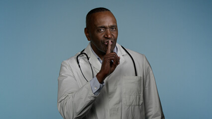 African American man doctor in blue studio hold index finger near mouth male medic medical worker...