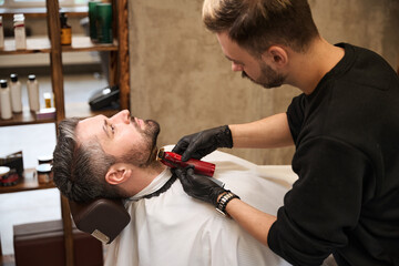 High-qualified barber trimming facial hair of handsome adult man