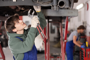 Young guy mechanic in uniform repairs underbody of car in car service station