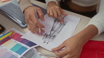 A man and a woman collaborate in a design studio, examining fashion sketches and fabric samples, with a laptop and colored pencils on desk