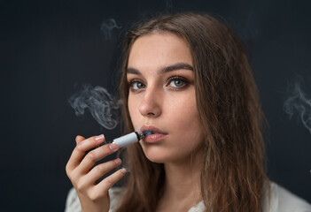 Fototapeta premium caucasian young woman vaping an E-Cigarette, portrait, close-up face and dark background, smoking, smoke around her, age 15-25