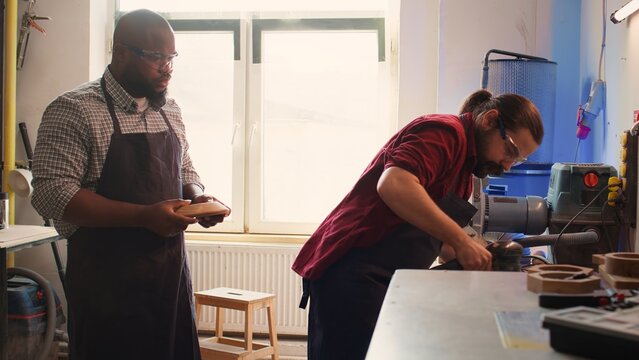Carpenter making modifications on blueprint drawings, using angle grinder on wood. Man using orbital sander gear after consulting schematics, assisted by african american apprentice, camera B