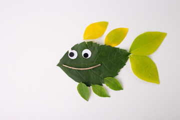 Fish made from leaves and natural materials. Body crafted from large green leaf, fins from smaller yellow and green leaves. Googly eyes and curved twig smiling mouth, everyday natural elements,
