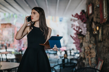 Businesswoman in elegant black dress talking on phone and holding tablet in stylish cafe. Focused professional in a busy environment.