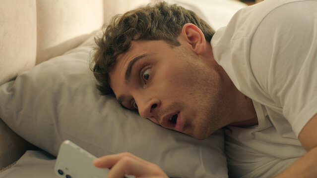 Man sleeping looking at screen mobile phone smartphone telephone late overslept male guy bedroom bed worried deadline morning routine stressed shocked rushing clock hurry punctuality panic quick fast