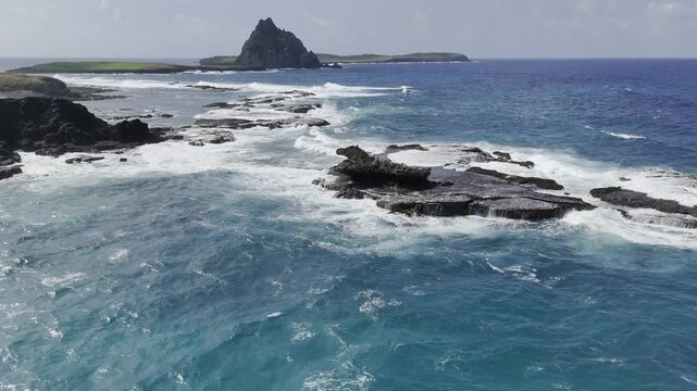 Drone flies right, low over waves crashing on rocks, with a looming Ilha Sela Gineta in the background in Fernando de Noronha, Pernambuco, Brazil