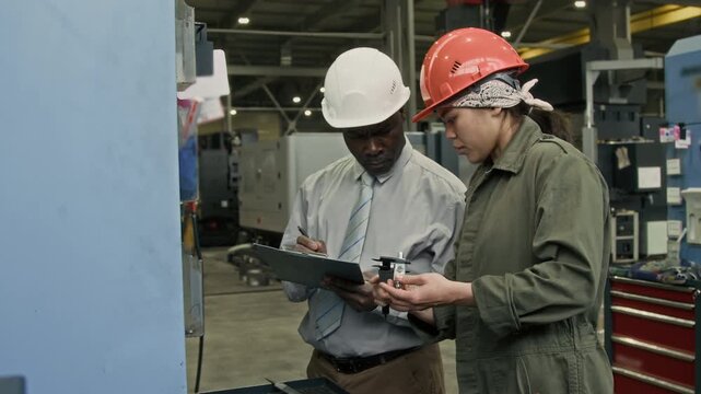 Medium shot of female Hispanic engineer scaling metal detail with vernier caliper and telling results to male Black supervisor who writing data into report while controlling output quality in plant