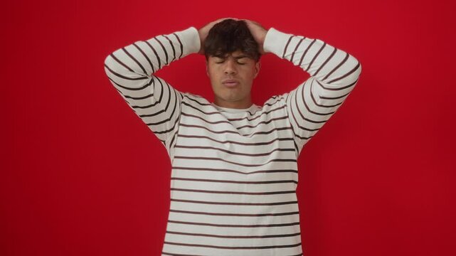 Manic moment, young hispanic man wearing stripes t shirt standing, head in crazy hands, scared and shocked with mouth open in surprise over isolated red background